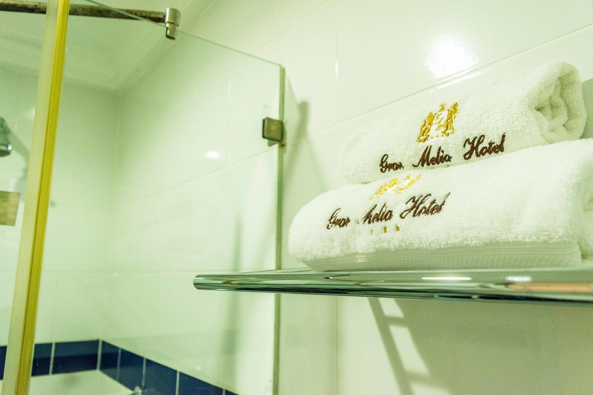A bathroom with a glass shelf holding a folded towel with "Grand Media Hotel" embroidered in gold.