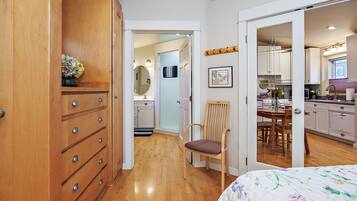 A bedroom with a wooden wardrobe, a bed with floral bedding, a chair, and a view into the kitchen.