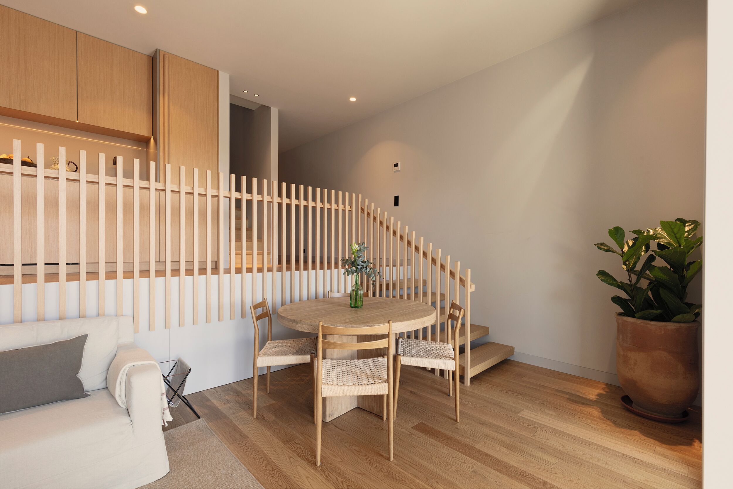 Un espacio de estar moderno con una mesa de comedor redonda de madera rodeada por cuatro sillas, un sofá blanco con cojines y una planta grande en maceta.