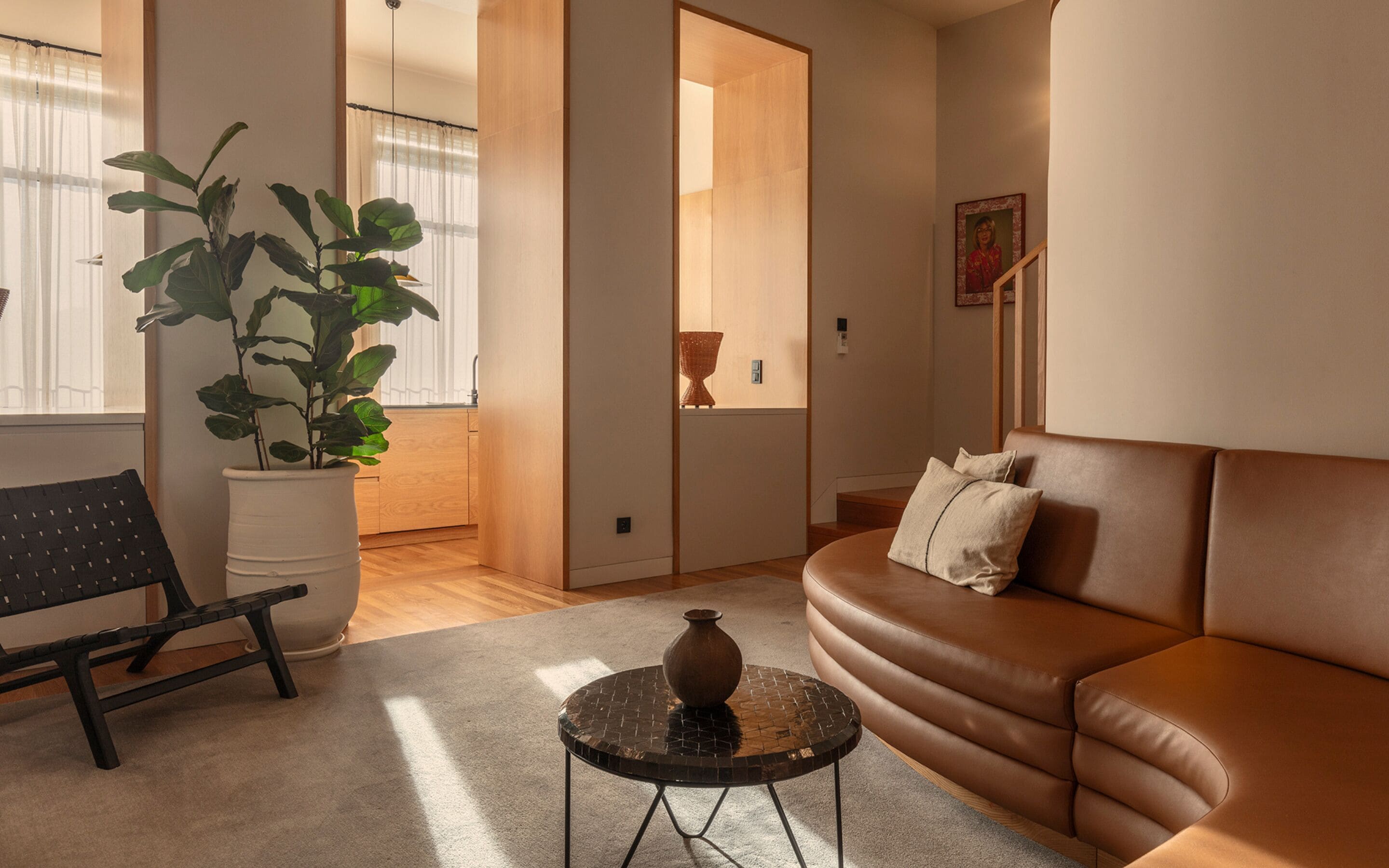 Una sala de estar con un sofá de cuero marrón, un banco tejido negro, una planta en maceta y una pequeña mesa redonda con un florero.