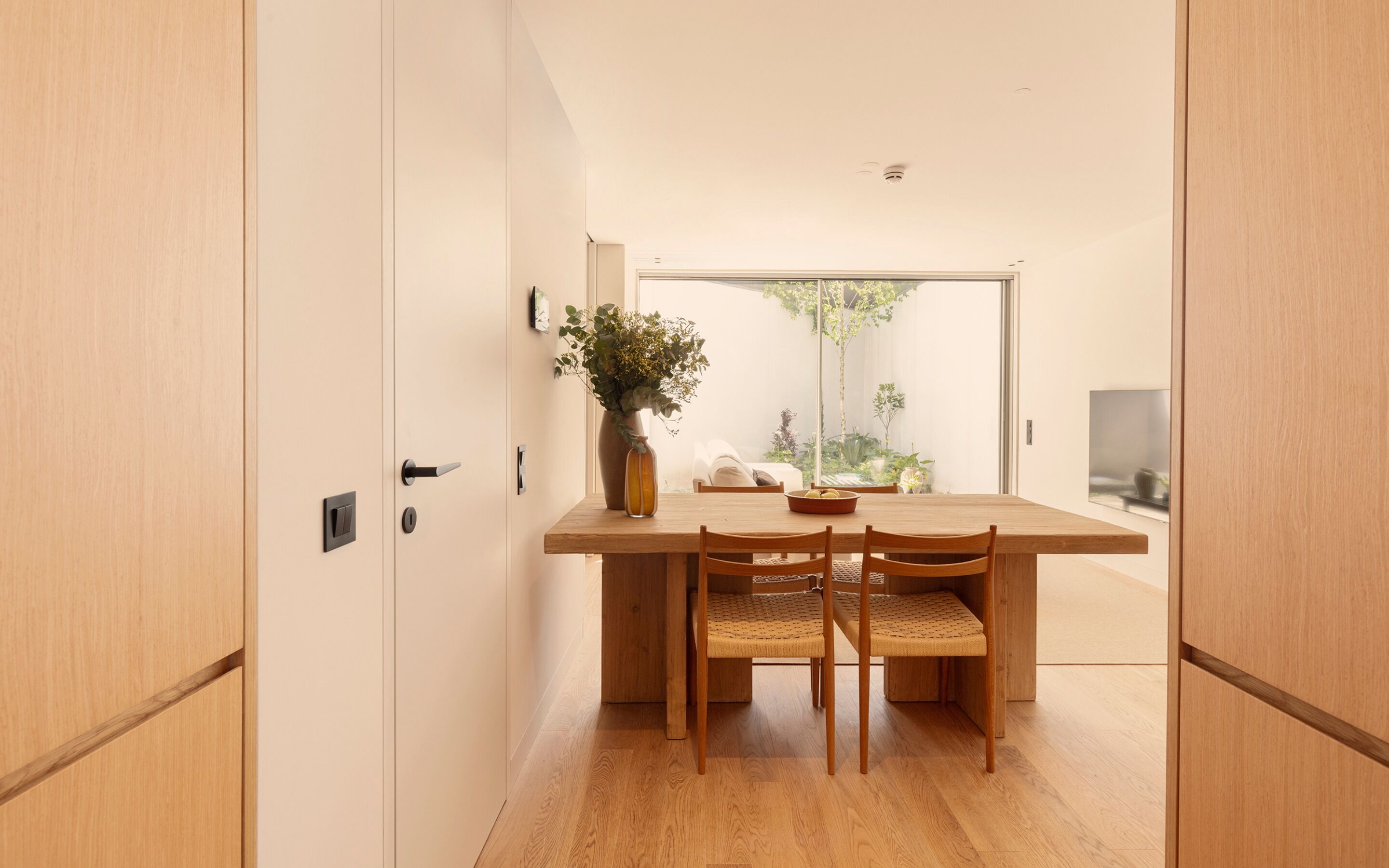 Comedor minimalista con una mesa y sillas de madera, un florero con flores y vista a zonas verdes a través de una puerta corrediza de vidrio.