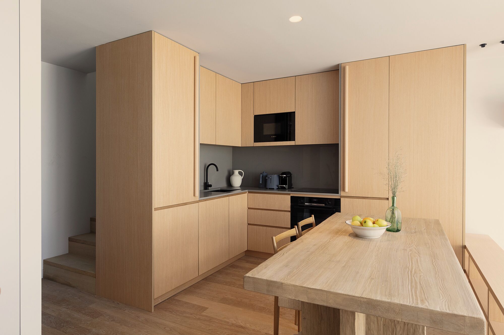 Cocina moderna con armarios de madera, microondas negro, un lavamanos y una mesa de comedor con un bol de fruta.