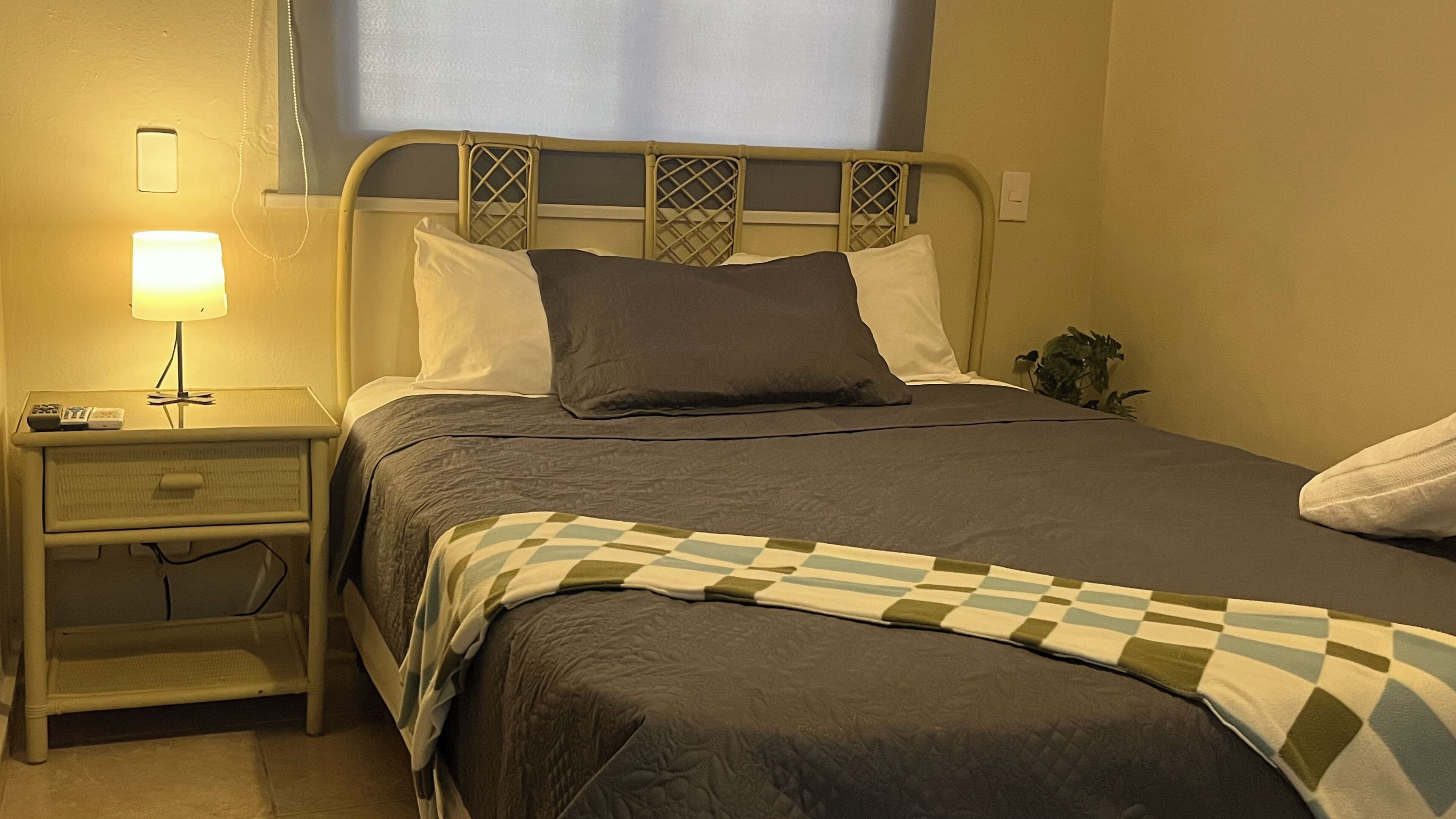 Una cama bien tendida con un edredón a cuadros, una lámpara de noche y una mesita de noche con un control remoto.