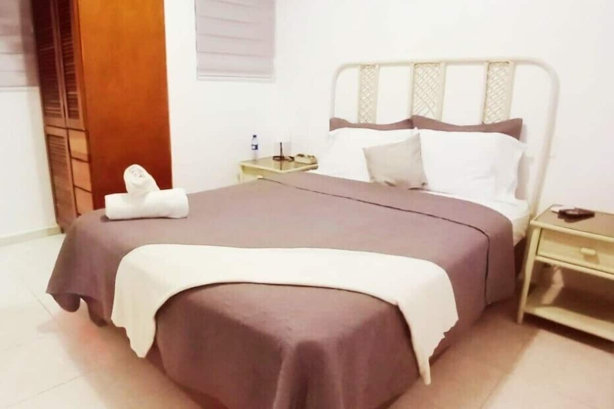 A neatly made bed with a brown bedspread and white pillows, a bedside table with a lamp, and a wooden door in the background.