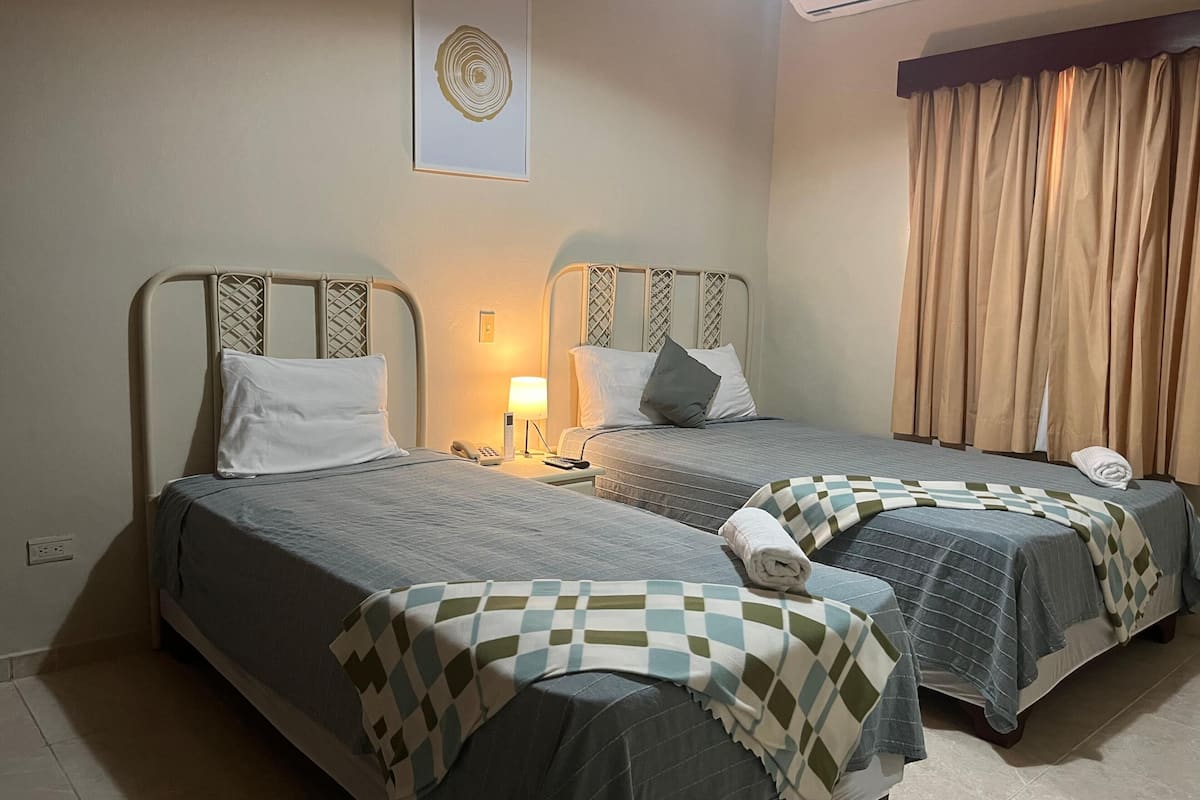 A hotel room with two beds, a bedside table with a lamp, a phone, and a wall-mounted air conditioner.