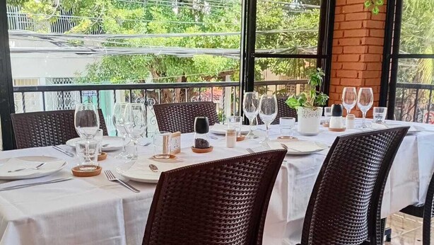 Comedor con una mesa dispuesta para cuatro personas, rodeada de sillas de mimbre, y vista a zonas verdes.
