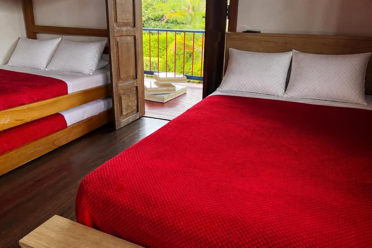 A hotel room with two beds, each with a red blanket and white pillows, wooden headboards, and a balcony with a view of greenery.