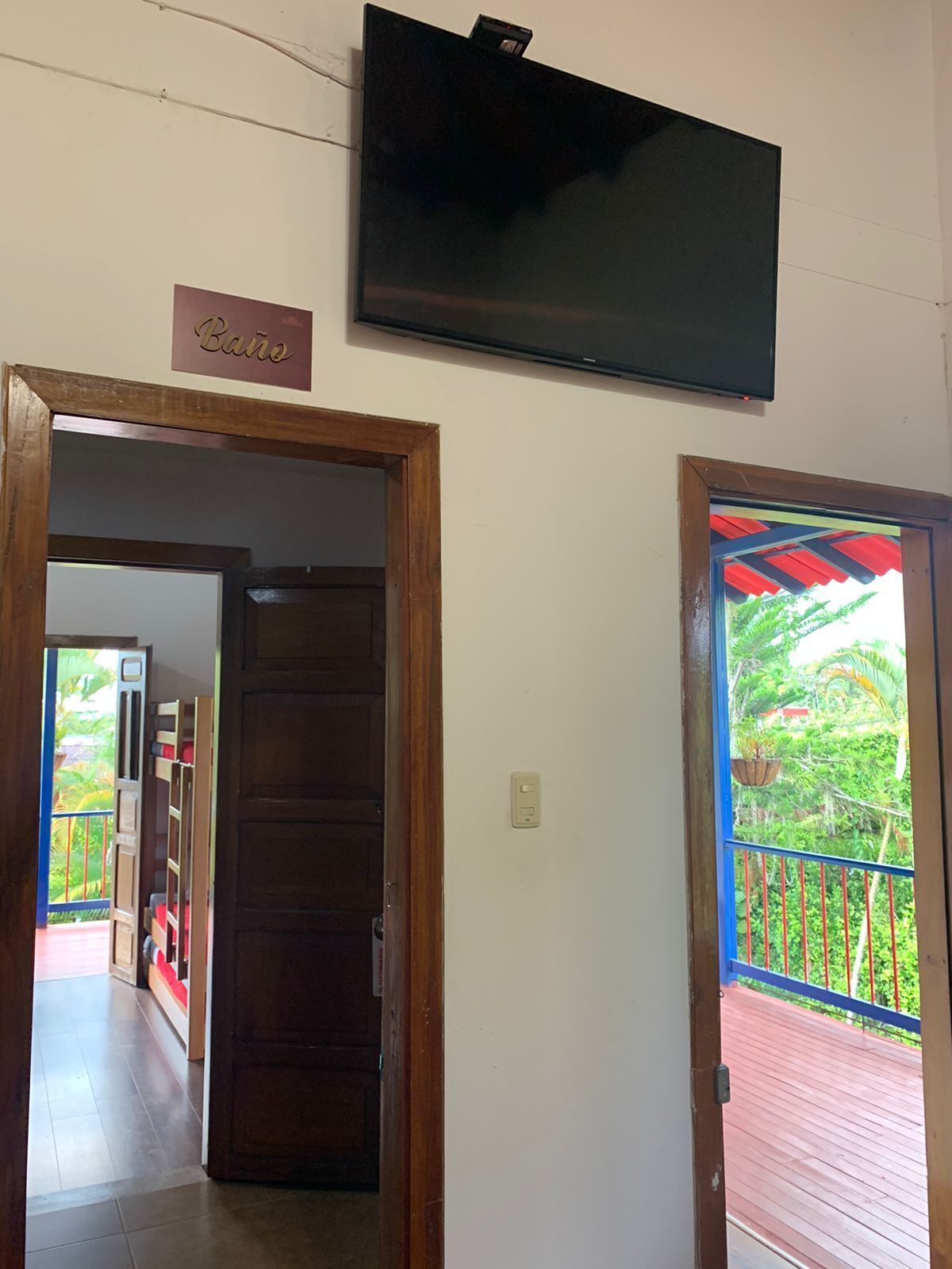 Quarto com porta de madeira, televisão fixada na parede e varanda com guarda-corpo vermelho.