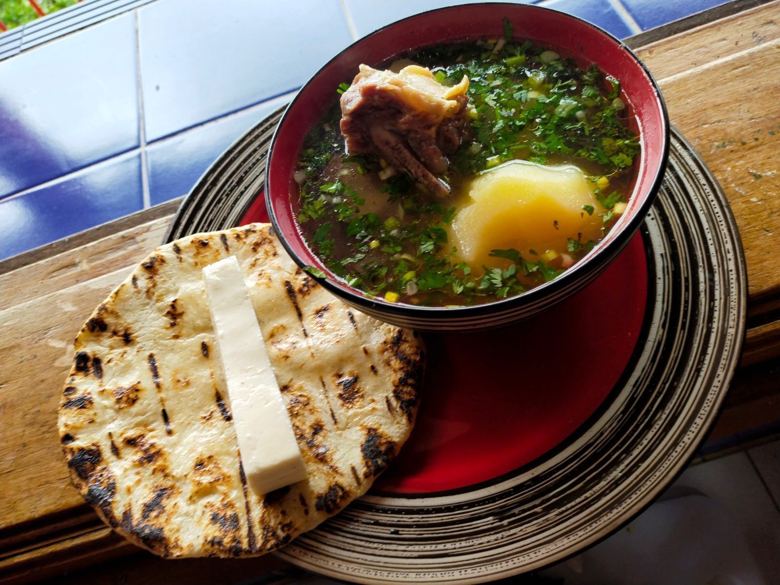 A bowl of soup with meat and vegetables, a piece of flatbread, and a red plate on a wooden table.
