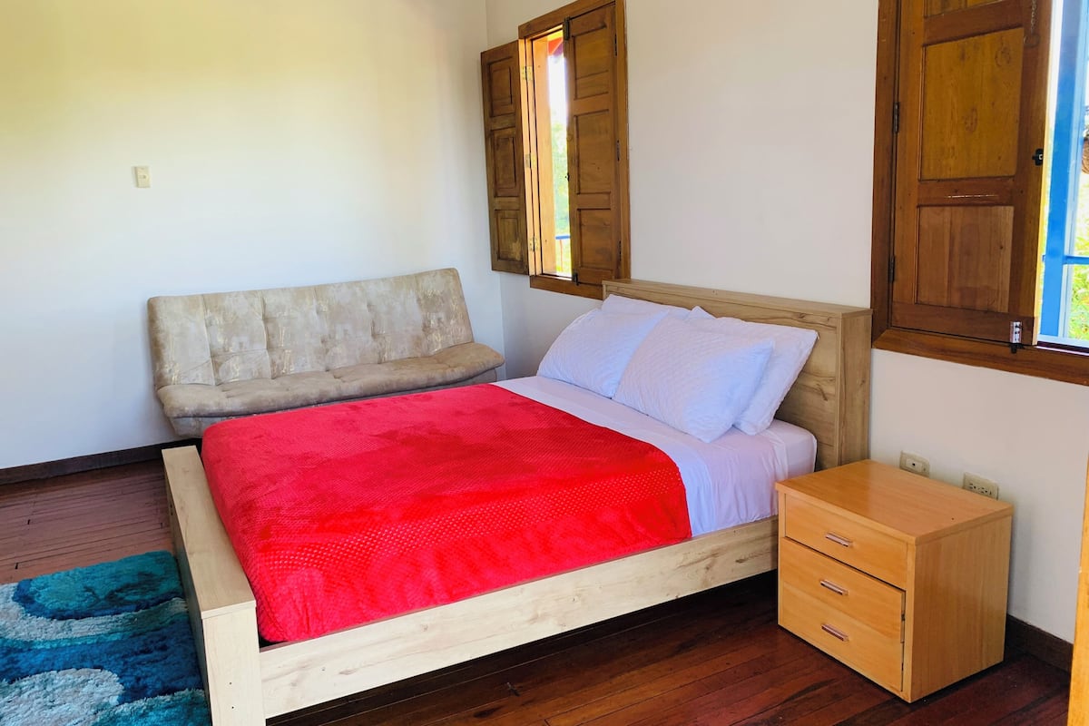 A bedroom with a wooden bed, a red blanket, a wooden nightstand, and a blue rug.