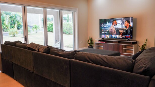 Ein Wohnzimmer mit einem großen Flachbildfernseher auf einem Schrank, einem braunen Ecksofa und einer Glastür, die in einen Garten führt.