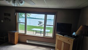 A room with a large window overlooking a lake, a television, a heater, and a small table.