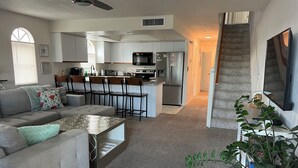 A modern living room with a grey sofa, a coffee table, and a dining area with bar stools. A kitchen with white cabinets and appliances is visible in the background. - Sun Kiss Villas - Beachside Duplex, Private Pool and Steps to Beach (Fort Myers Beach)