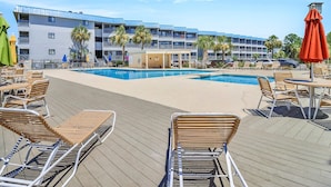 A poolside area with lounge chairs and umbrellas, a pool, and a multi-story building with balconies in the background. - Sun, Sand, Serenity -Savannah/Tybee (Tybee Island)