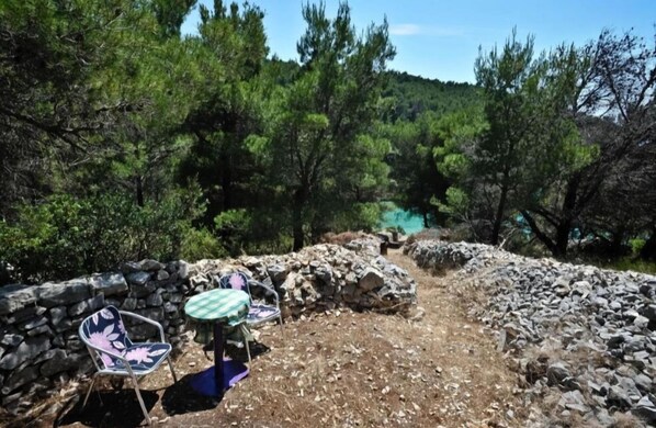 A rustic outdoor seating area with stone walls and two chairs, surrounded by dense greenery and a clear view of a turquoise body of water. - Seclude charmy house with pool 200m from sea (Duboka)