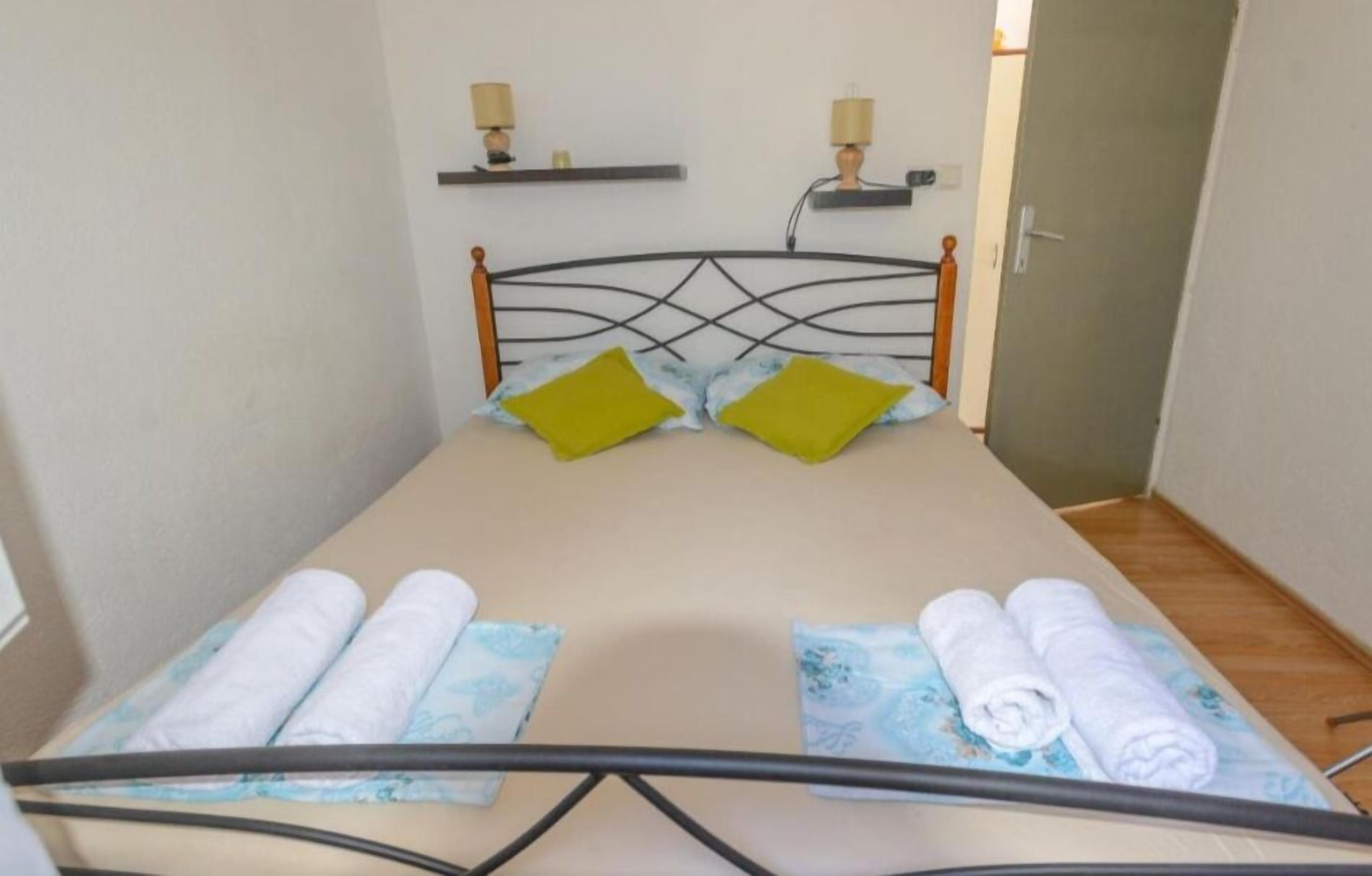 A small, neatly made bed with a metal frame, two pillows, and two rolled towels on a patterned bedspread.