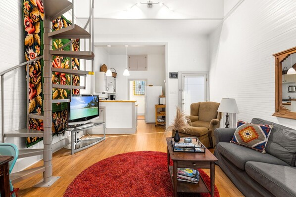 Una sala de estar moderna con alfombra roja, sofá gris, televisor y escalera con arte mural colorido.