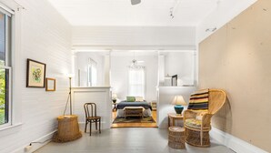 A spacious room with a large window, a bed with a blue bedspread, wicker chairs, and a red rug.