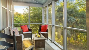 A covered porch with wicker furniture and a table, large windows, and a view of trees.