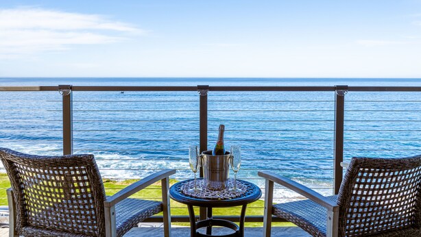 Un balcon avec des fauteuils en osier et une table sur laquelle se trouvent une bouteille de champagne et des verres, offrant une vue sur l’océan.