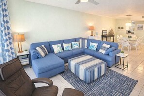 A living room with a blue sectional sofa, a patterned ottoman, a coffee table, a side table with a lamp, and a dining area with white chairs and a table.