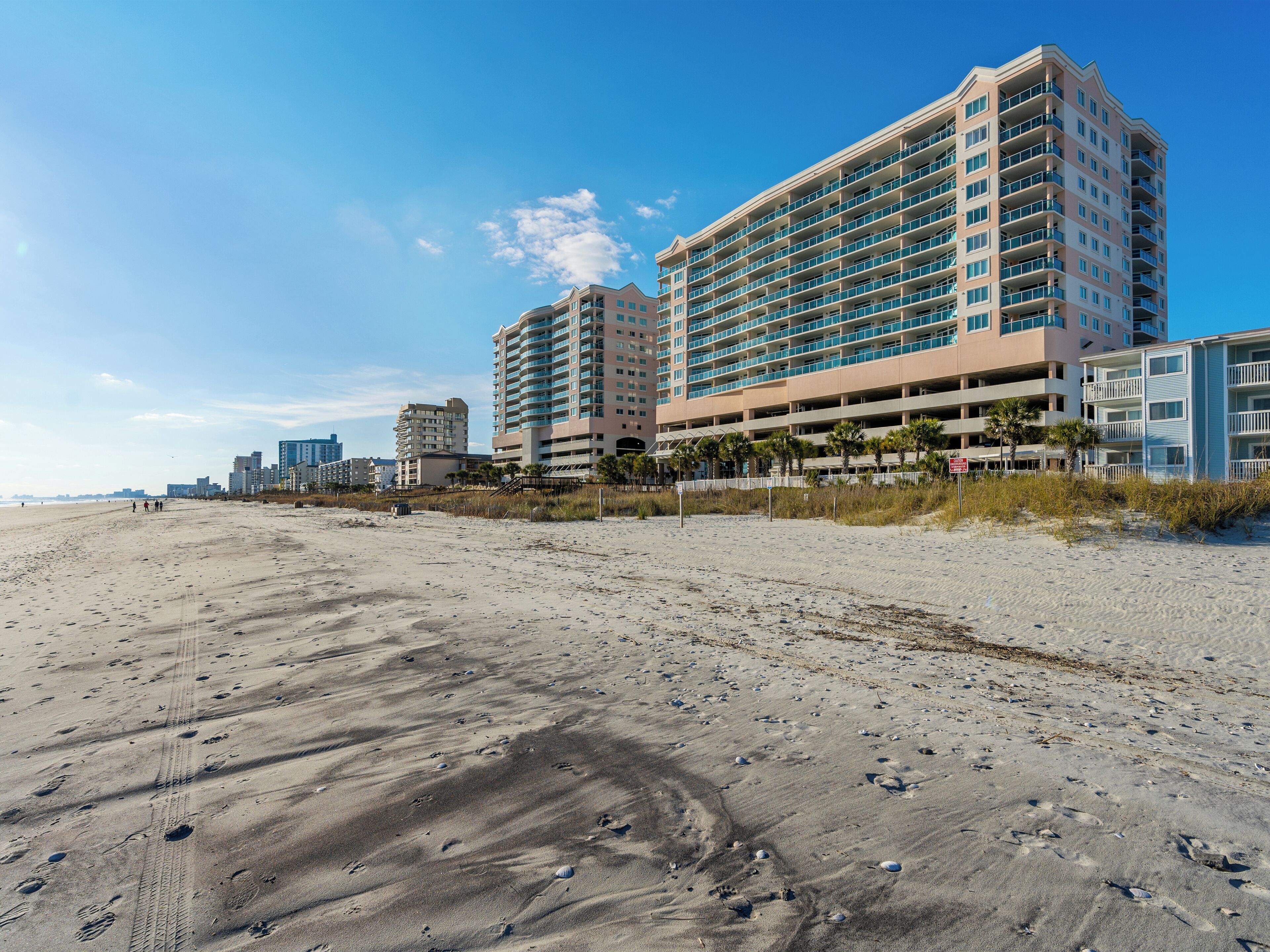 Een hotel aan het strand met meerdere verdiepingen, een zandstrand op de voorgrond en een heldere lucht.