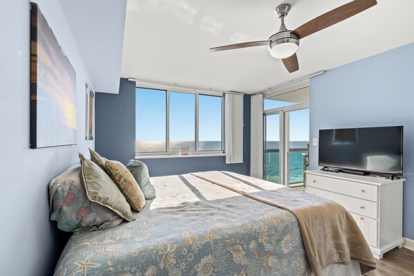 Une chambre à coucher avec un lit, une télévision, un ventilateur de plafond et une grande fenêtre donnant sur la mer.