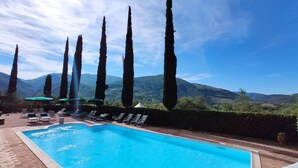 Piscina com espreguiçadeiras e guarda-sóis, cercada por árvores altas e montanhas ao fundo.