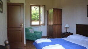 Quarto com cama, cadeira verde, guarda-roupa de madeira e janela com vista para o verde.