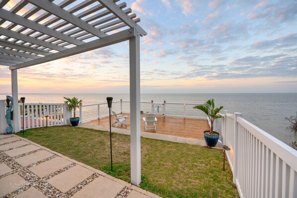 Una terraza techada con barandilla que da a un cuerpo de agua, con pérgola, plantas en macetas y mobiliario exterior.
