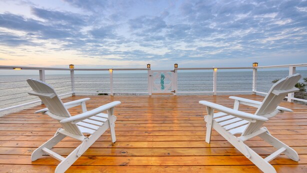 A wooden deck with two white Adirondack chairs facing a body of water under a cloudy sky. - Bungalow on South Padre Bay (Port Isabel)