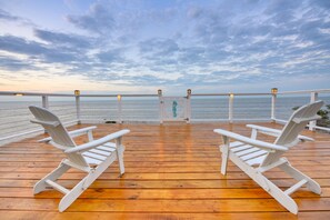 Una terraza de madera con dos sillones Adirondack blancos mirando un cuerpo de agua bajo un cielo nublado.