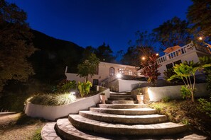 A nighttime view of a building with a stone staircase leading up to a balcony, surrounded by greenery and illuminated by ambient lighting.