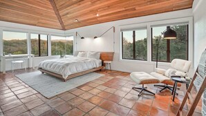 A spacious bedroom with a large bed, a desk, and a chair, large windows, and a view of the outdoors.