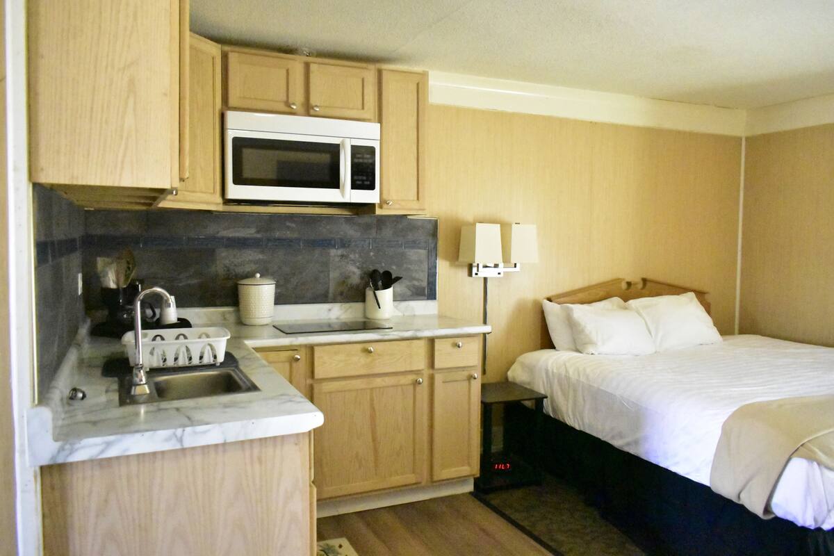 A compact room with a kitchenette, a bed, and a nightstand.