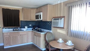 A compact kitchen with wooden cabinets, a microwave, and an air conditioning unit. There is a small dining area with a table and chairs. - Hampton Motel (Hampton)