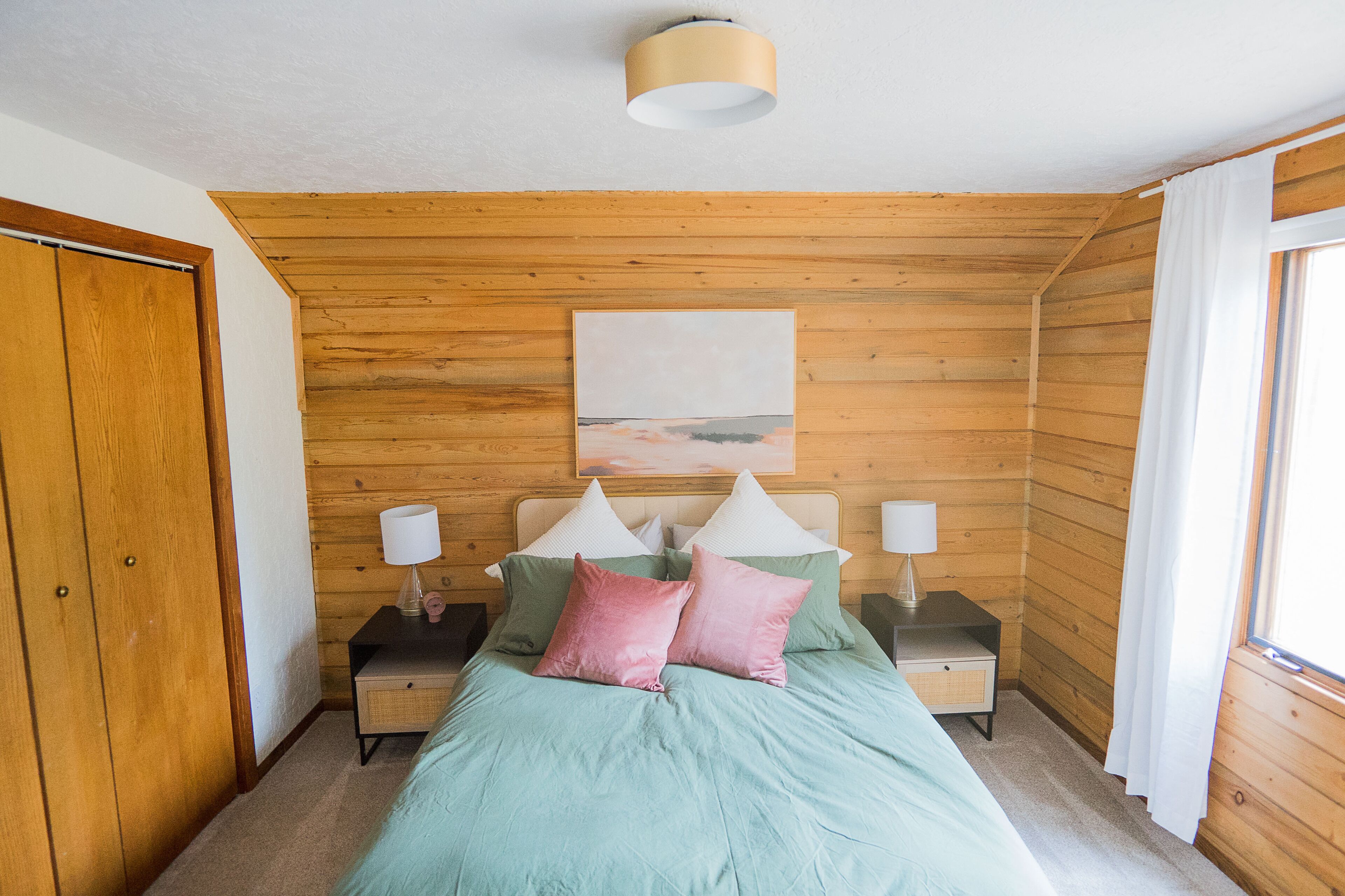 A bedroom with wooden walls, a bed with green and pink bedding, two bedside tables with lamps, and a painting on the wall.