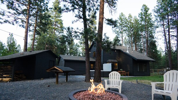 A fire pit with flames in a circular metal container, surrounded by gravel. There are two white Adirondack chairs and a wooden bench nearby. - Family-Friendly, Modern Home with Extra Large Hot Tub with EV Charger (Sisters)
