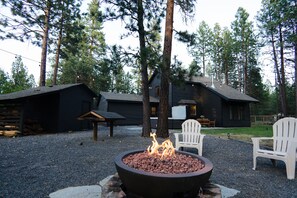 A fire pit with flames in a circular metal container, surrounded by gravel. There are two white Adirondack chairs and a wooden bench nearby. - Family-Friendly, Modern Home with Extra Large Hot Tub with EV Charger (Sisters)