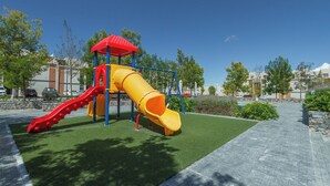 Parquinho infantil com um escorrega vermelho e amarelo, cercado por grama sintética verde e um calçadão pavimentado.