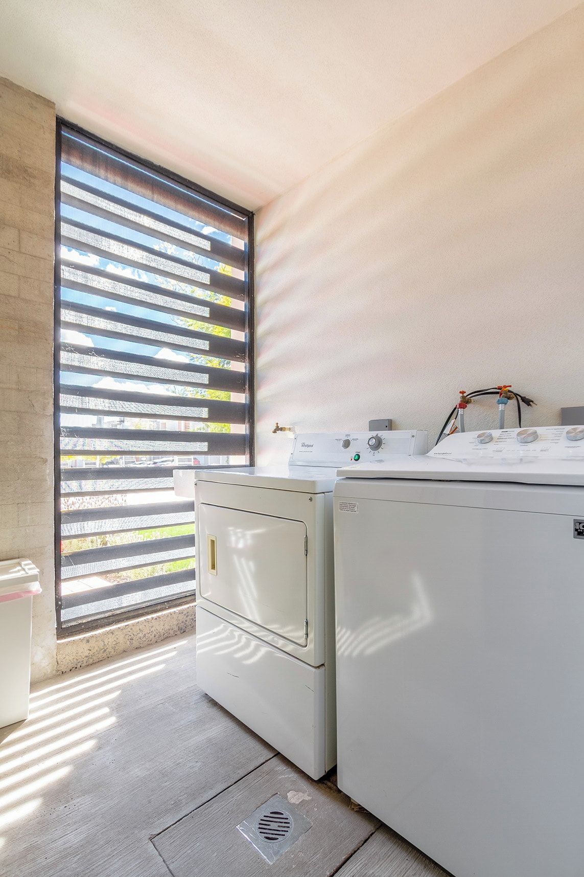 Une buanderie équipée de deux machines à laver et deux sèche-linge blancs, une fenêtre avec des stores et un sol en béton.