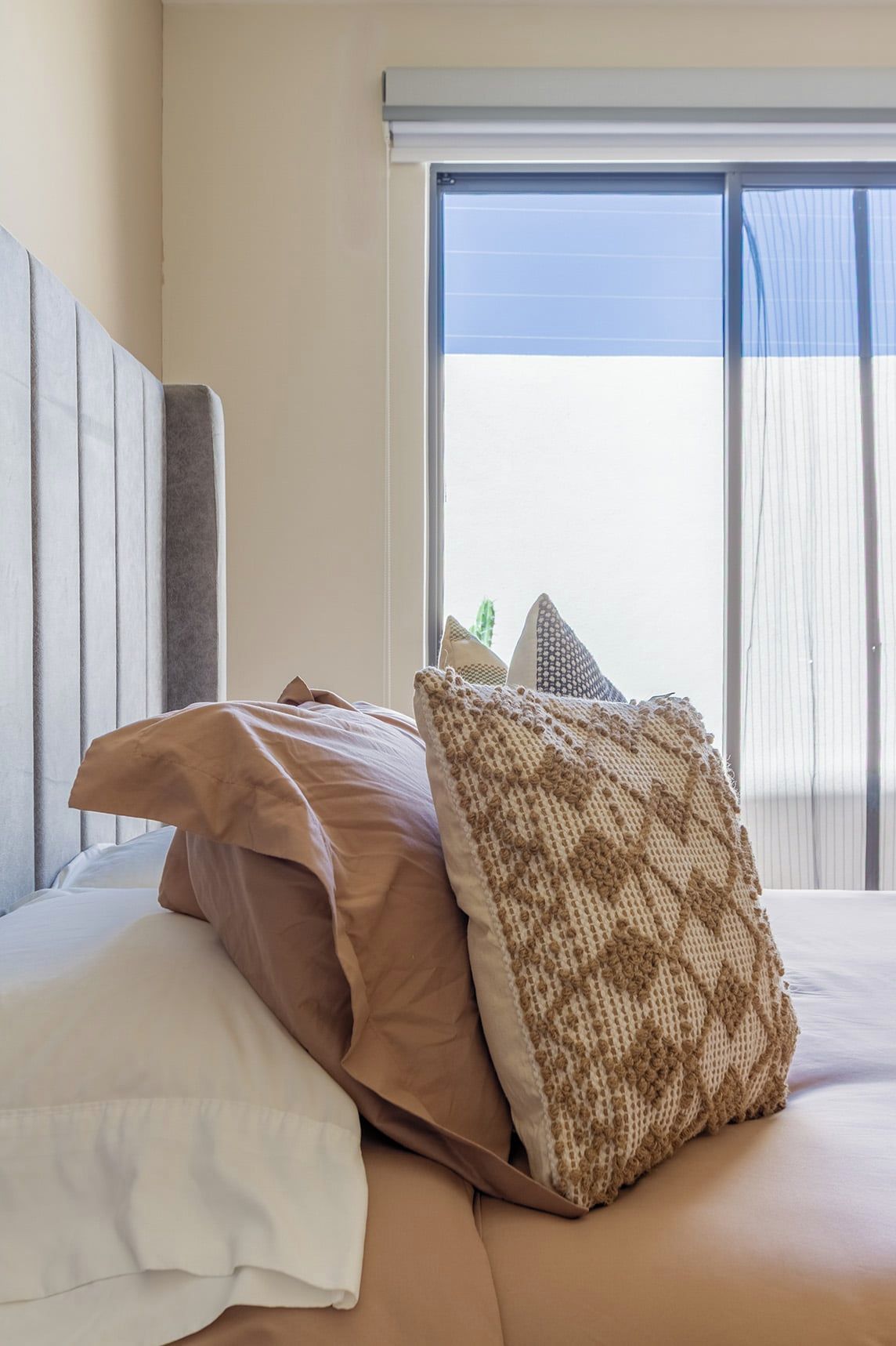 A bed with a beige mattress, a white pillow, and a brown knitted pillow.
