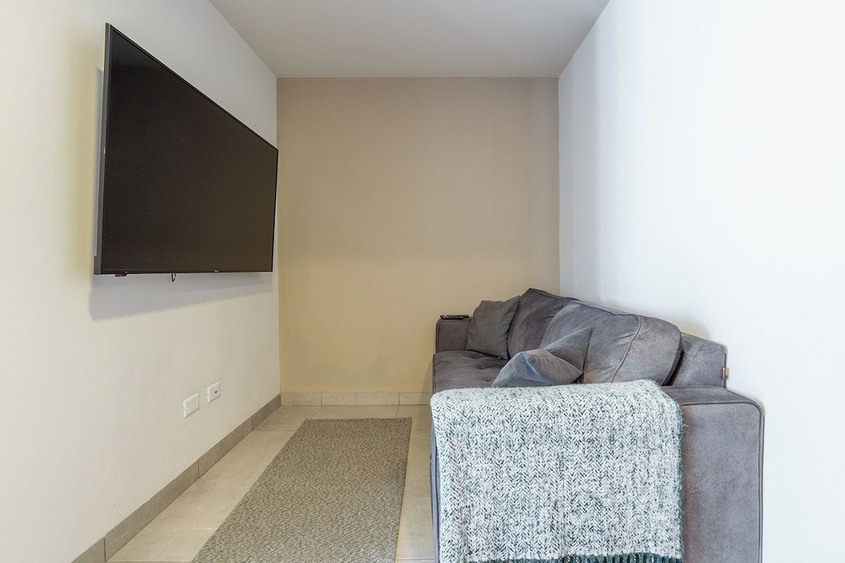 A living room with a flat-screen TV mounted on the wall, a grey sofa, and a grey rug.