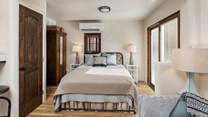 Un dormitorio estrecho y bien iluminado con una cama grande, dos mesitas de noche con lámparas, un armario de madera y una ventana con persianas.