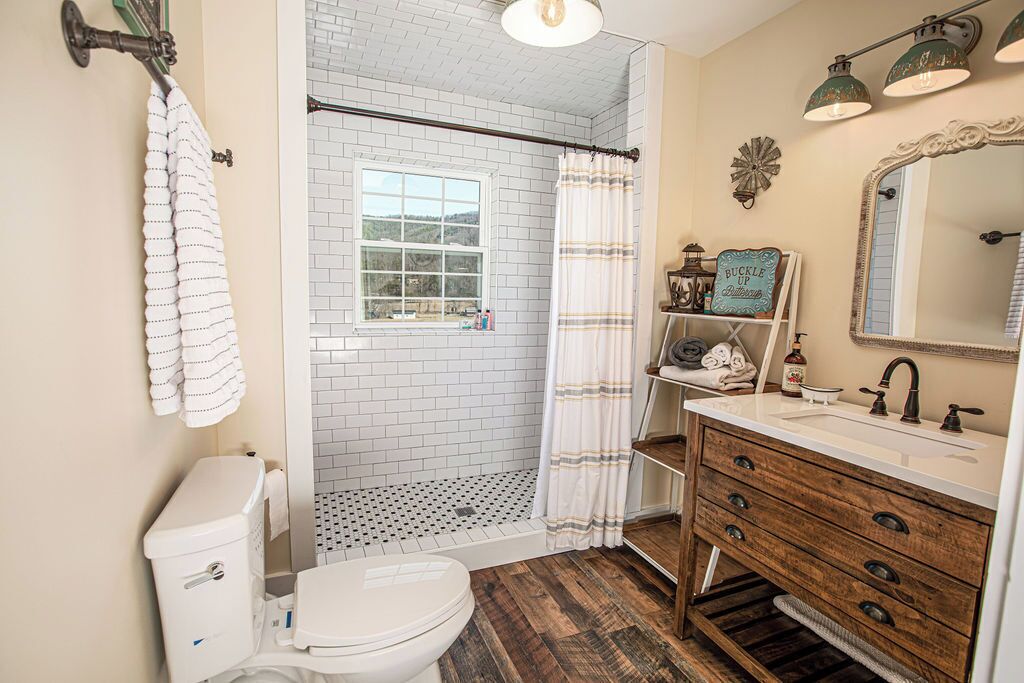 Une salle de bain avec des toilettes blanches, une douche avec un rideau blanc, un meuble-lavabo en bois avec un miroir et une fenêtre donnant sur l’extérieur.