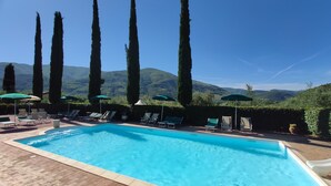 A swimming pool with lounge chairs and umbrellas, surrounded by tall trees and a mountainous backdrop. - COLLEOLIVO: Three-room apartment in farmhouse with swimming pool, Marmore Falls (Ferentillo)