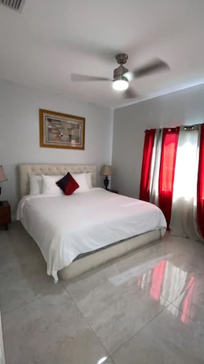 Un dormitorio con una cama grande, un ventilador de techo, una mesita de noche con lámpara y un cuadro en la pared.