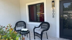 A small outdoor seating area with two wicker chairs and a table, a window with a curtain, and a door with a number sign.