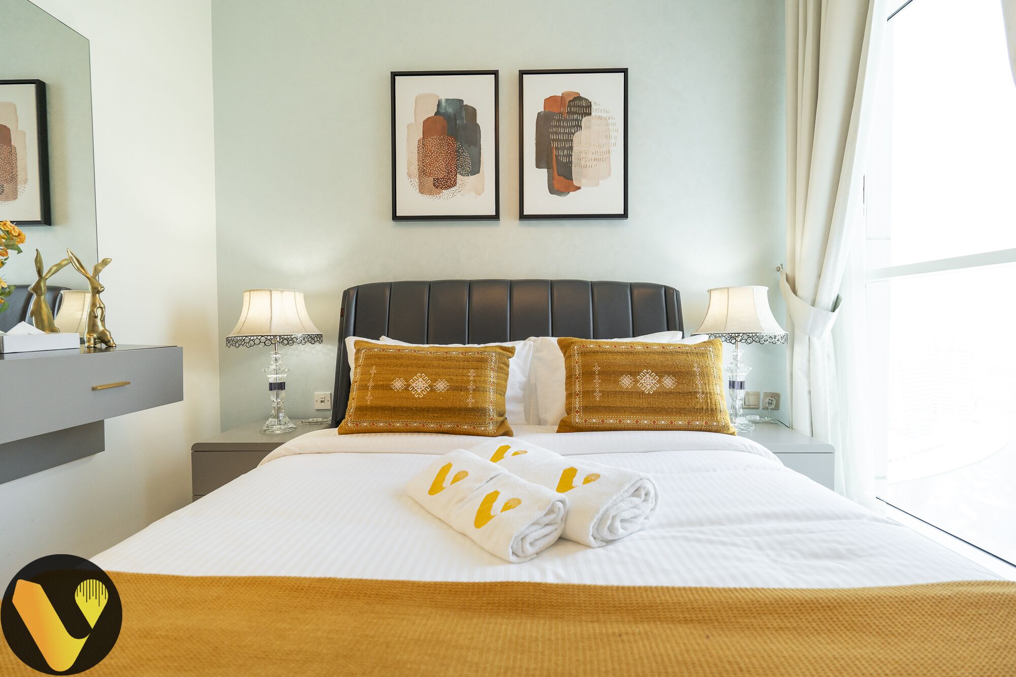 A hotel room with a bed, two pillows, a nightstand, a lamp, a mirror, and two framed artworks on the wall.
