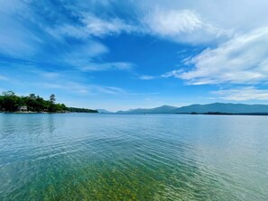 A calm lake with clear water, surrounded by trees and mountains under a blue sky with clouds.